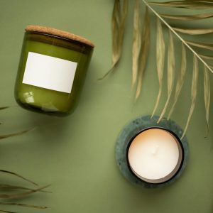 Aesthetic display of a green jar candle and a lit scented candle with natural leaves on a green background.