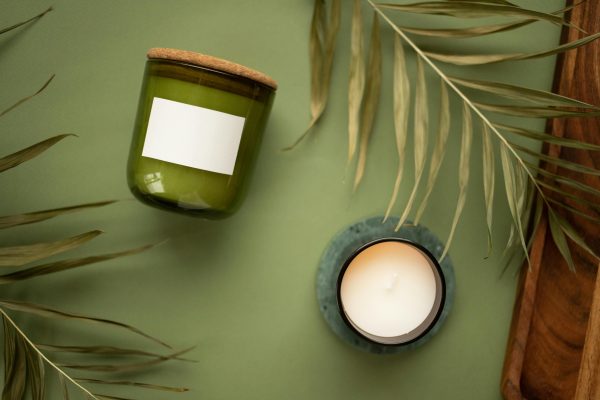 Aesthetic display of a green jar candle and a lit scented candle with natural leaves on a green background.